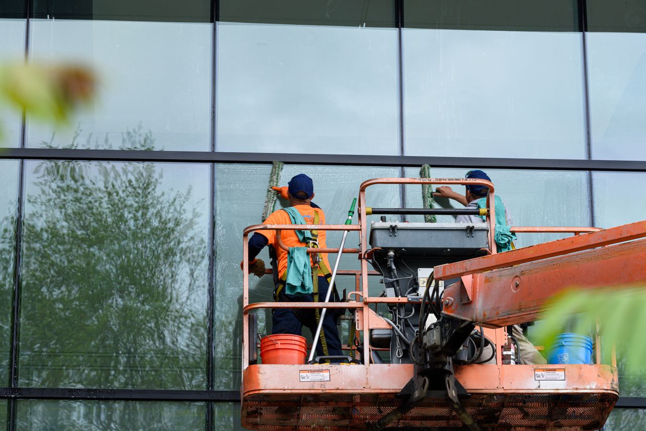 services-03 Two workers cleaning windows on a glass building facade using a lift platform, enhancing building aesthetics.