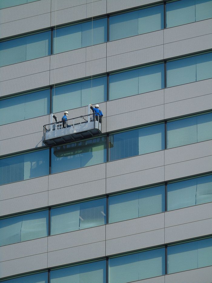 gallery-04 Two workers clean windows on a modern glass building facade using a suspended platform.