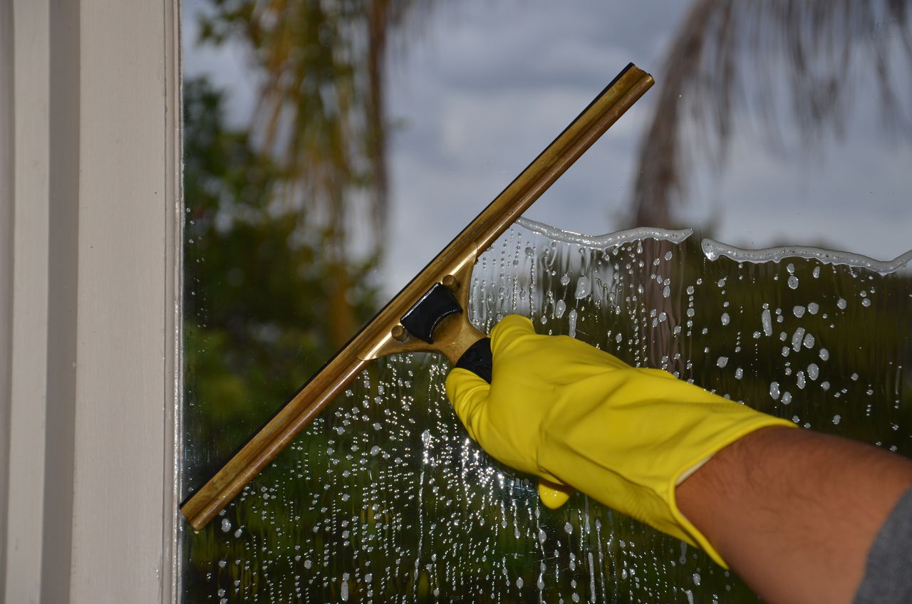 gallery-02 Close-up of hand cleaning window with squeegee outdoors using yellow gloves.